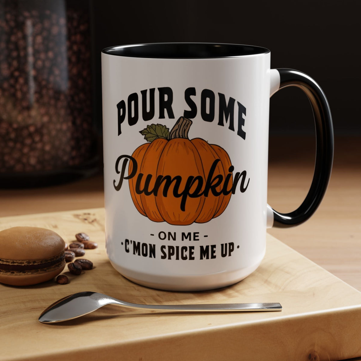A white ceramic mug with a black handle and interior, printed with the phrase 'Pour Some Pumpkin - On Me - C'mon Spice Me Up' and an orange pumpkin illustration. The mug sits on a wooden board with a spoon and coffee beans nearby.