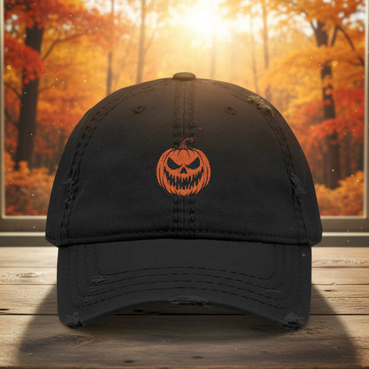 A black distressed baseball cap with an orange embroidered jack-o'-lantern on the front, displayed on a wooden surface with a blurred autumn forest background.