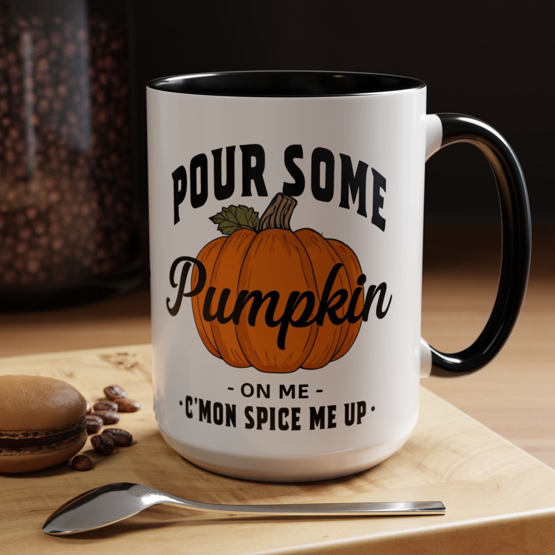 A white ceramic mug with a black handle and interior, printed with the phrase 'Pour Some Pumpkin - On Me - C'mon Spice Me Up' and an orange pumpkin illustration. The mug sits on a wooden board with a spoon and coffee beans nearby.